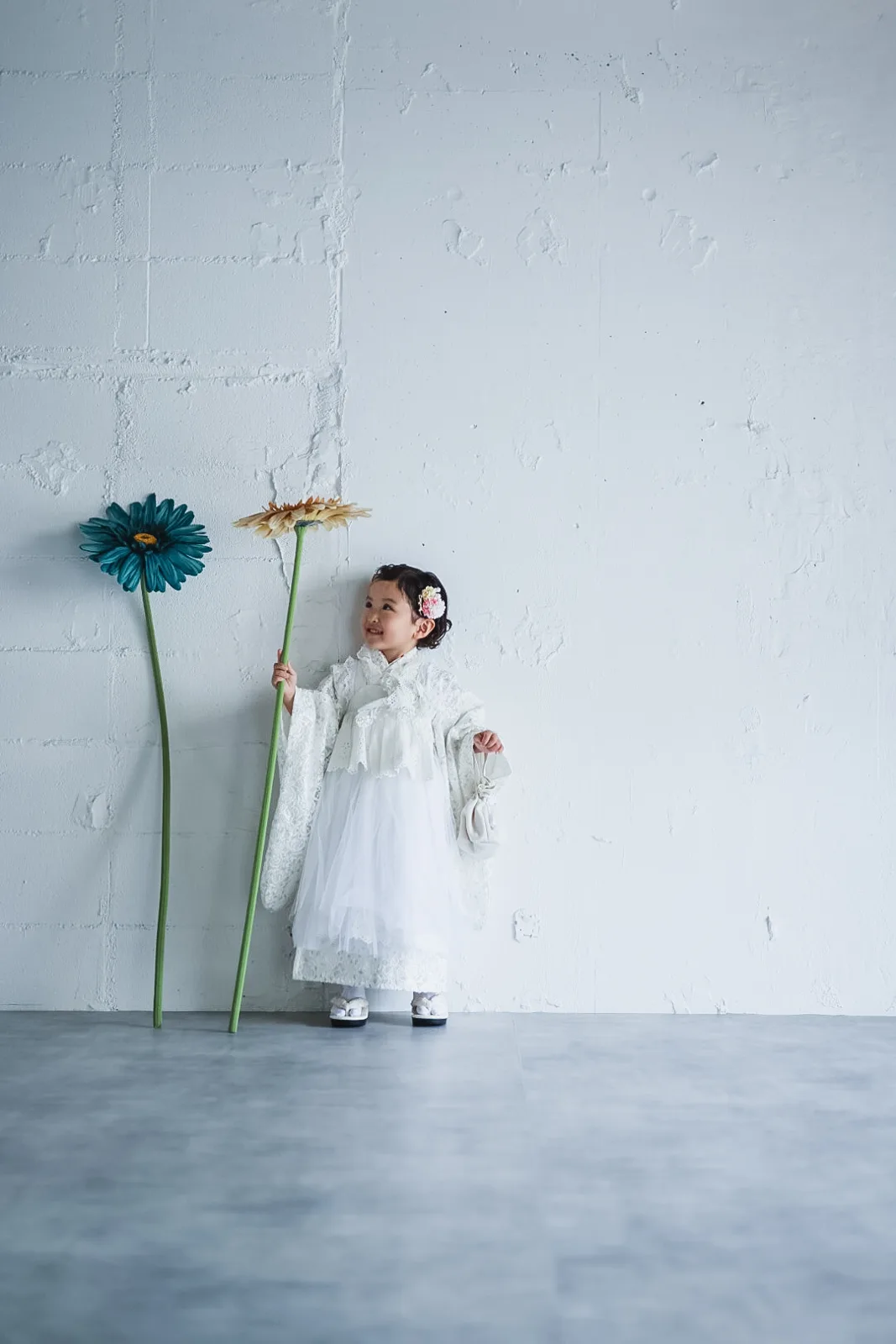 白い壁の前で、大きな花を持つ3歳の女の子